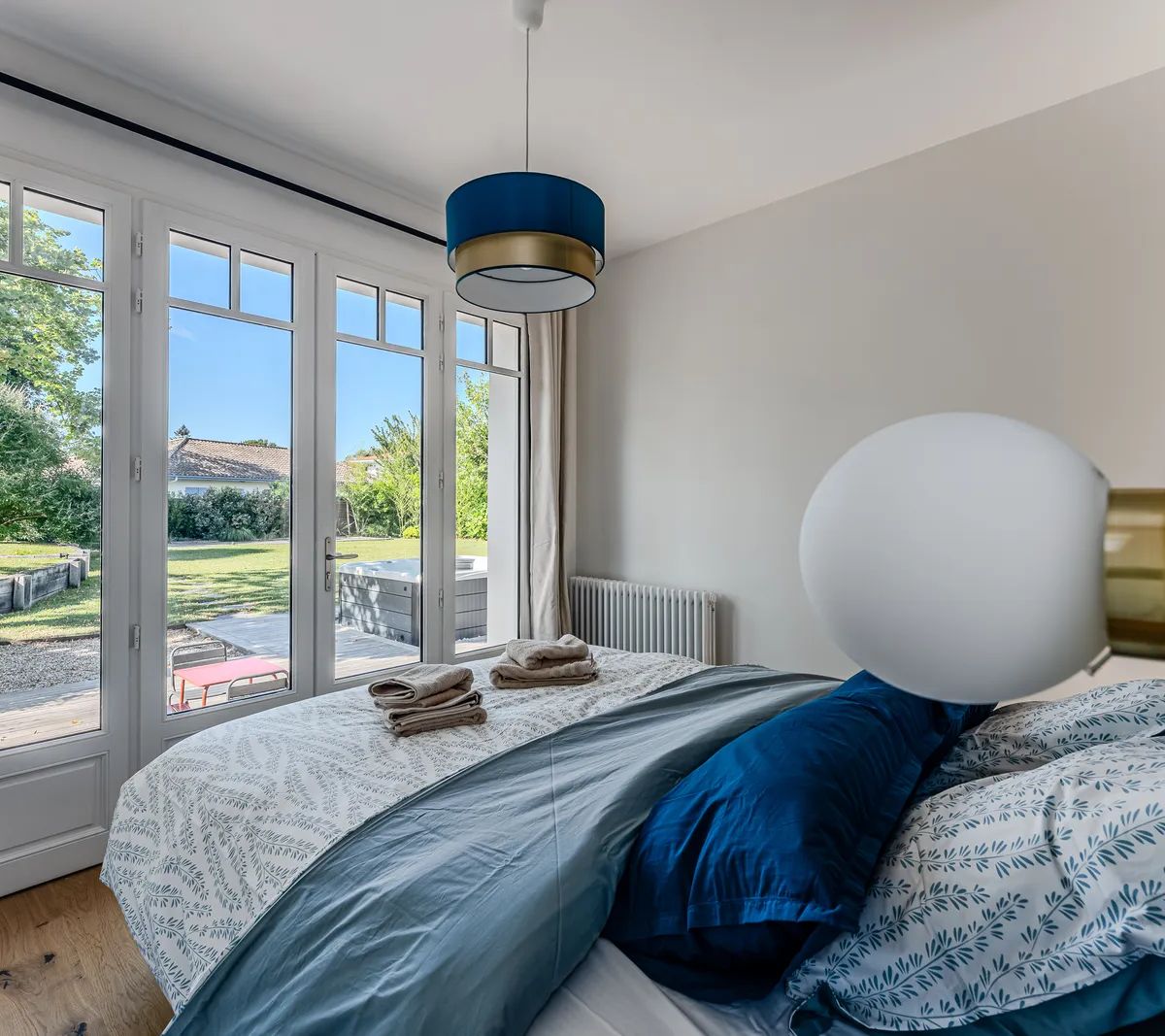 Chambre moderne avec grandes baies vitrées, suspension bleue et linge de lit coordonné donnant sur jardin