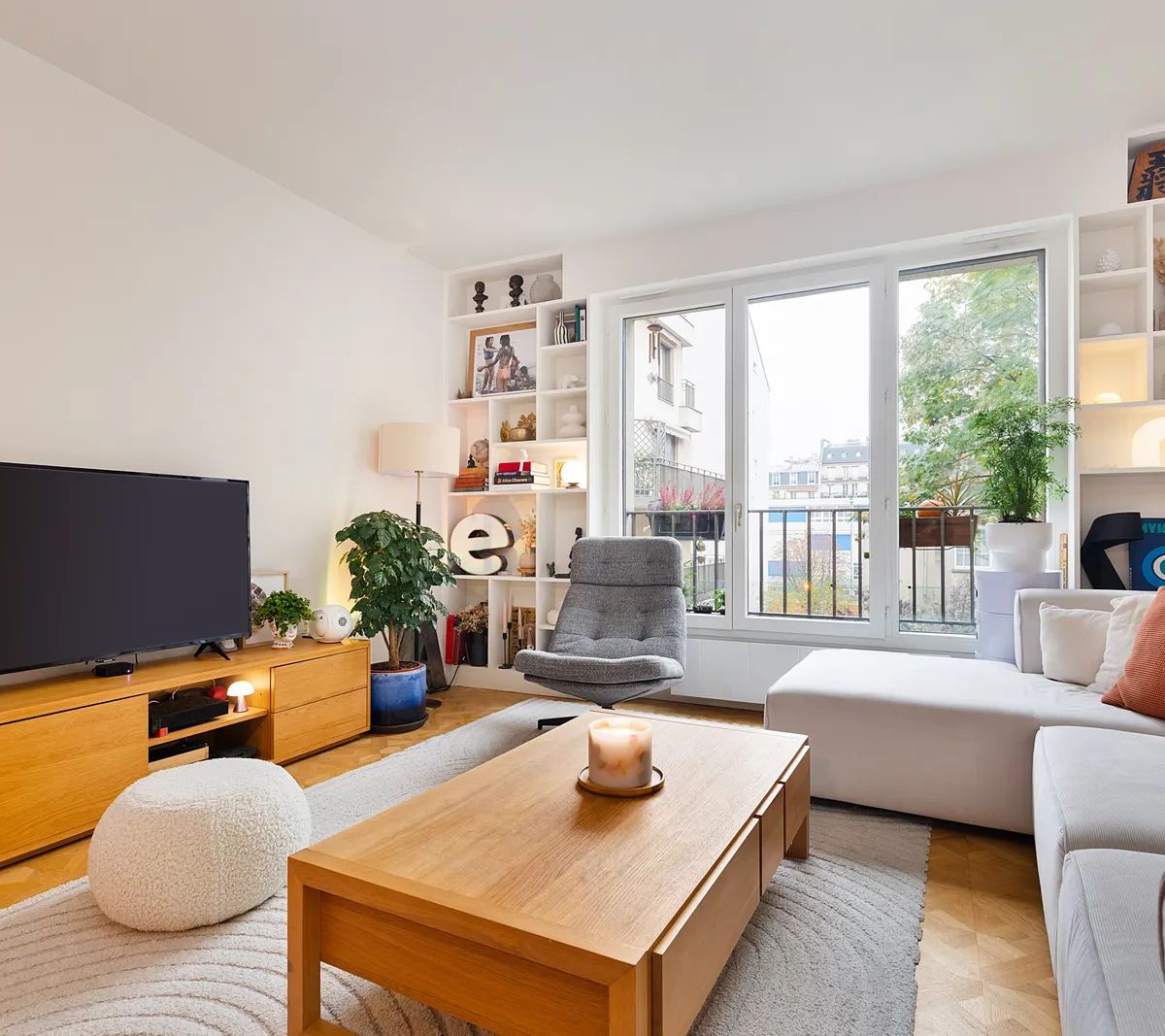 Salon moderne lumineux avec canapé gris, meuble TV bois, bibliothèque intégrée et grandes fenêtres donnant sur balcon
