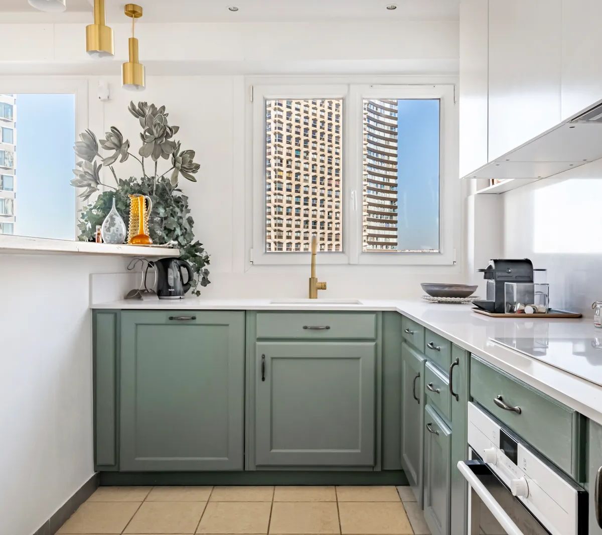 Cuisine moderne avec placards verts, plan de travail blanc et grandes fenêtres donnant sur immeubles urbains