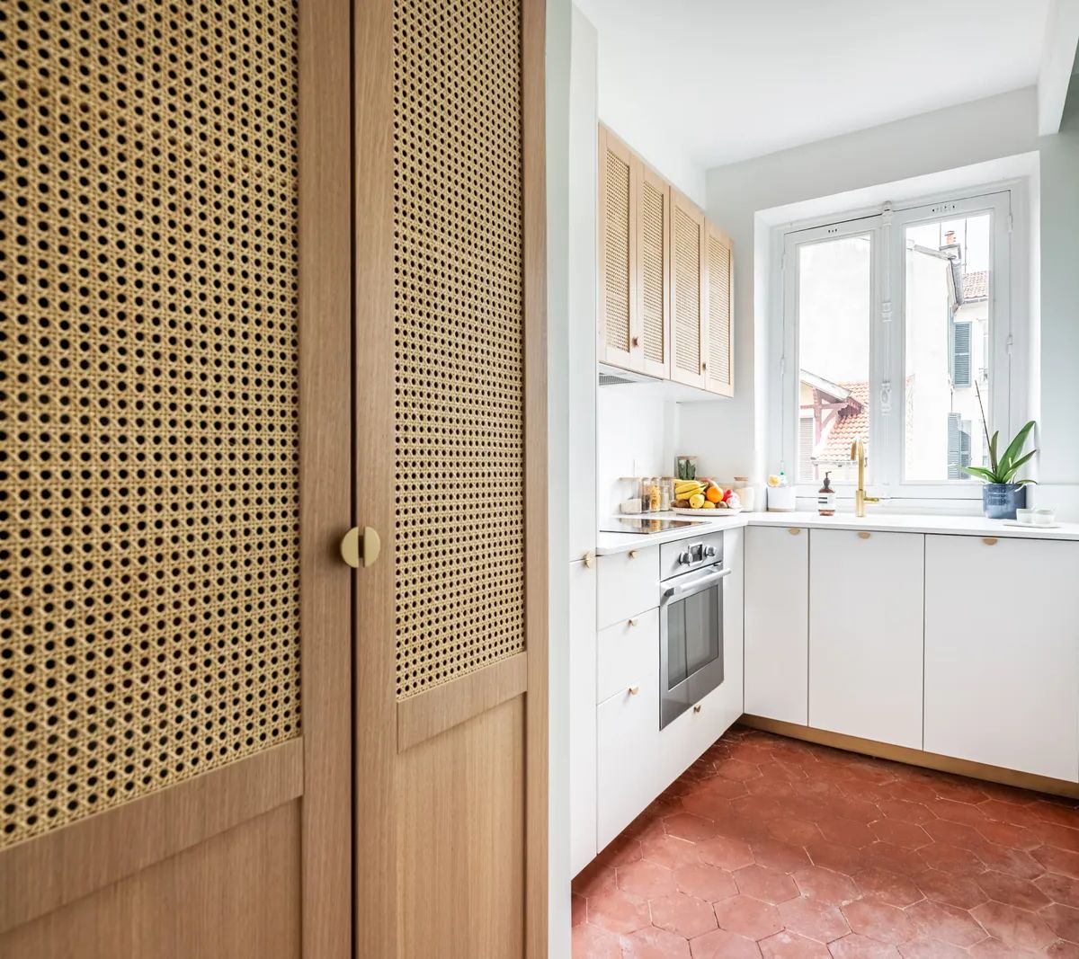 Cuisine moderne rénovée avec placards en cannage, sol en tomettes rouges et façades blanches dans appartement lumineux