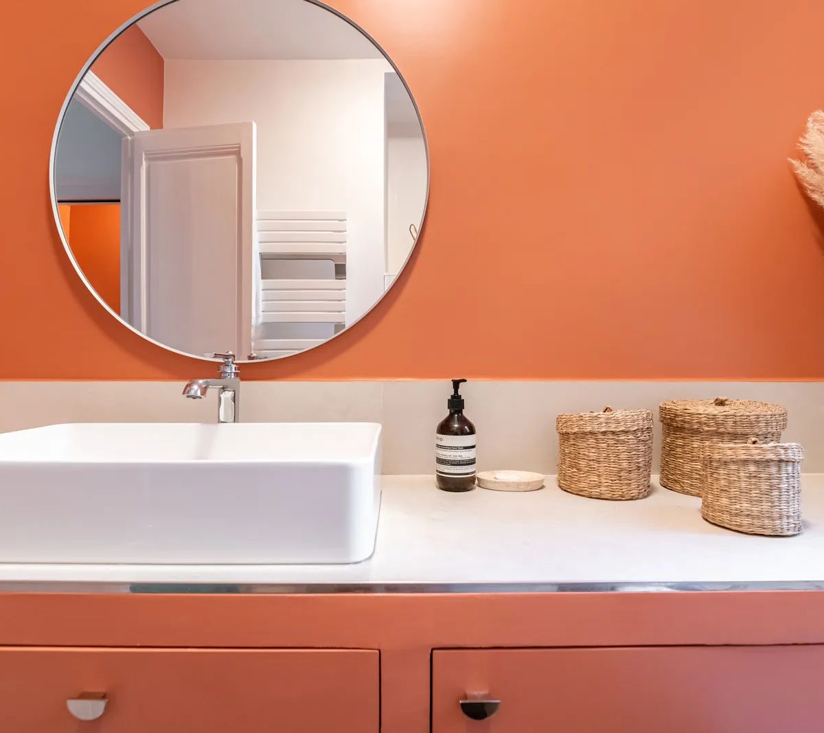 Salle de bain moderne avec mur orange, vasque blanche, miroir rond et rangements en osier sur meuble bois