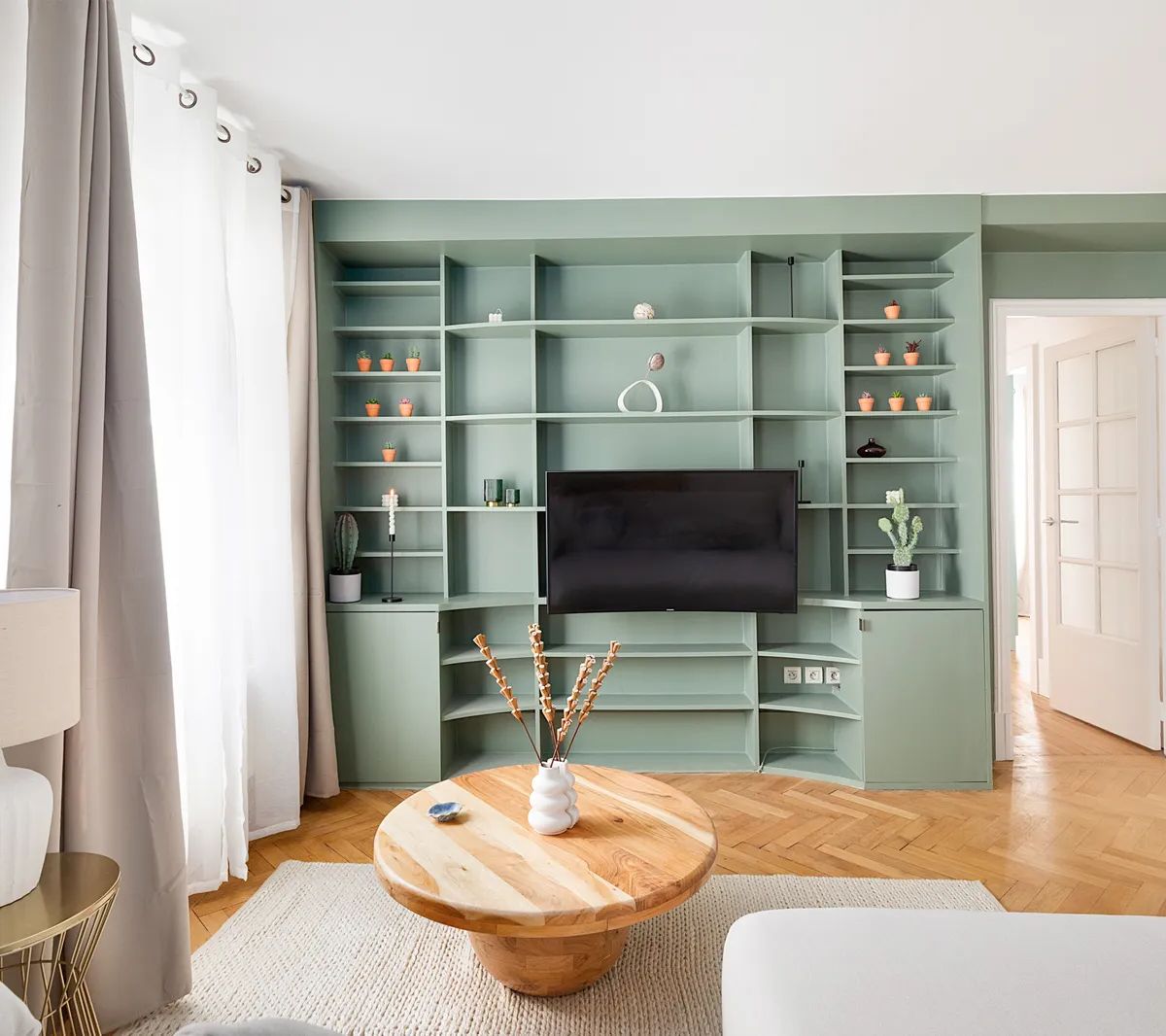 Salon moderne avec bibliothèque verte intégrée, parquet chevron, table basse ronde en bois et fauteuils blancs